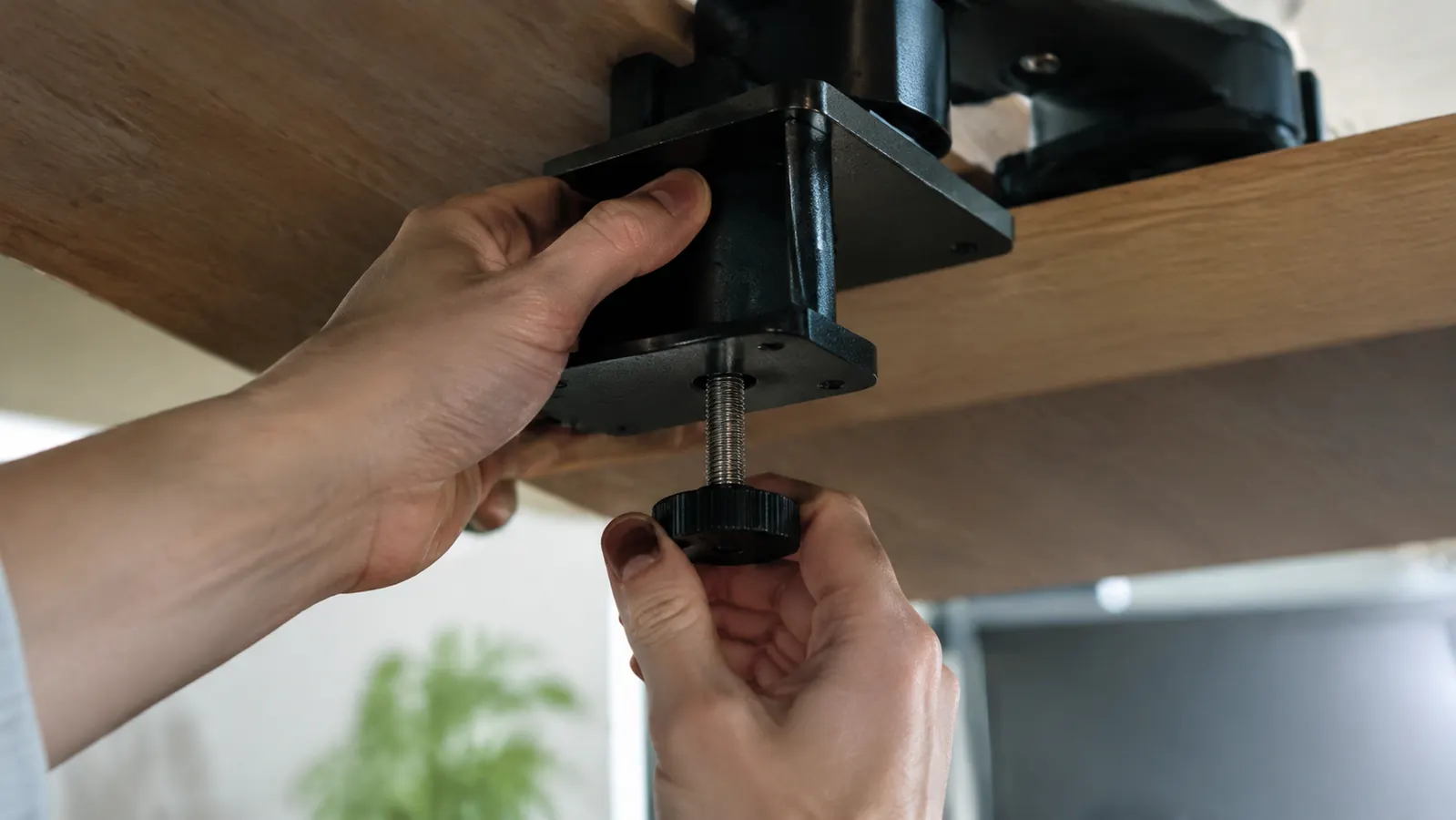 Hands beneath a desk tightening the clamp of a monitor mount