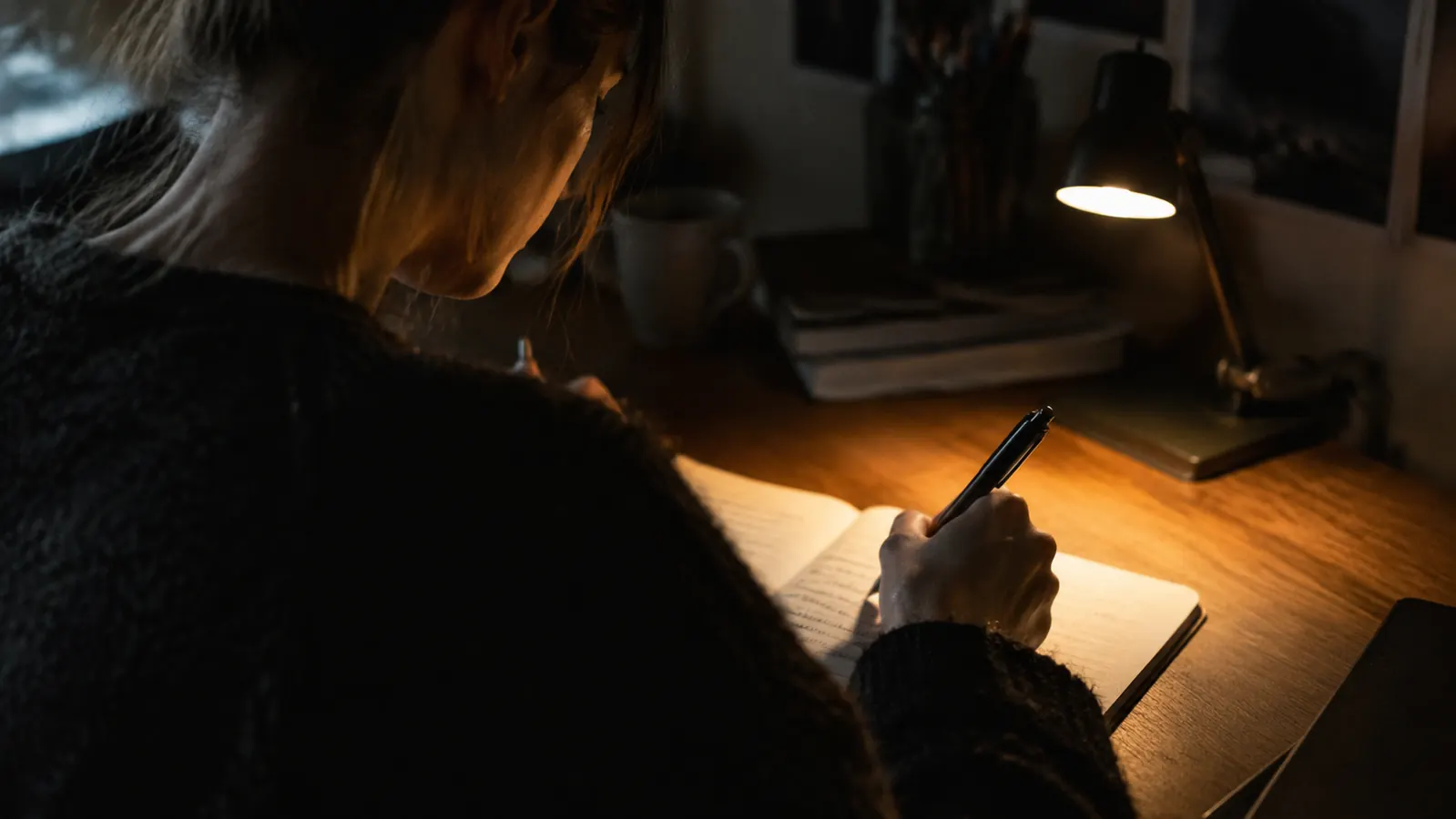 A person writing in a notebook at a desk lit only by a small lamp in the evening.