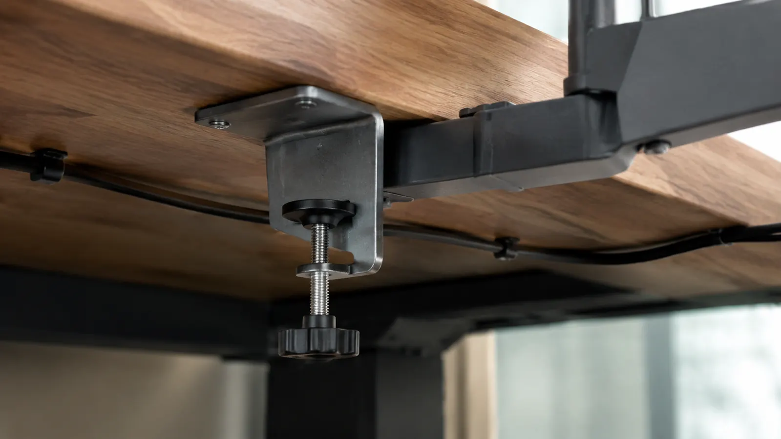 Underside of a desk showing a C-clamp base holding the monitor arm in place